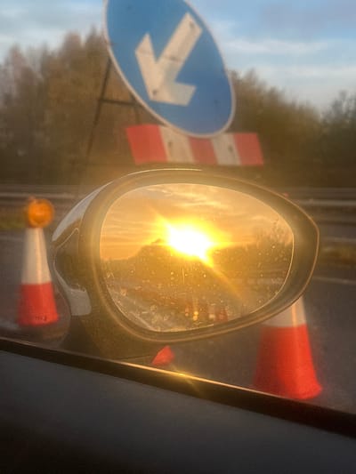 The image captures a close-up of a vehicle's passenger-side wing mirror, reflecting a brilliant, low-angle sun. The mirror surface exhibits numerous streaks and spots, interacting with the intense solar glare originating from the reflected sky. Outside the vehicle, to the left of the mirror, a white and orange traffic cone with an amber-colored beacon is partially visible. Another similar cone is partially visible to the right, behind the mirror. In the blurred background, a blue circular road sign with a white downward-pointing arrow indicates a mandatory direction, positioned above a red and white striped temporary barrier. Beyond these objects, a dense line of trees or foliage is discernible against a partly cloudy sky. The scene depicts a roadside environment with visible roadworks or traffic management apparatus, located in North Warwickshire, United Kingdom. No people are present. The interior door panel of the vehicle is visible at the bottom of the frame.