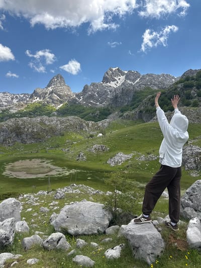 A person in a white hooded sweatshirt and brown pants is standing on a large rock outcropping. The person's arms are outstretched. The background features a landscape of grassy hills, rocky terrain, and mountains with patches of snow. In the distance, power lines run across a meadow-like area that could be prone to flooding based on ground texture and coloration. The scene is Brskut, Montenegro.