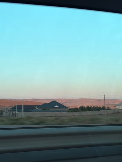 The image depicts a landscape scene from Kydyr Mambetuly, Kazakhstan. The view is seen through a window, with a dark, vertical frame on the left and vertical blinds on the right. The sky transitions from a pale blue to a soft yellow or peach hue, suggesting either dawn or dusk. In the background, rolling hills or mountains with reddish-brown soil are visible.

In the mid-ground, a modern-looking building with a dark, triangular roof is situated. Adjacent to it, there are grassy areas and some trees. A utility pole with wires stretches across the scene. The overall impression is one of a rural or semi-rural setting, likely in a region with arid or semi-arid terrain. The lighting suggests a calm and serene time of day. No people are visible in the frame, and there is no visible text.