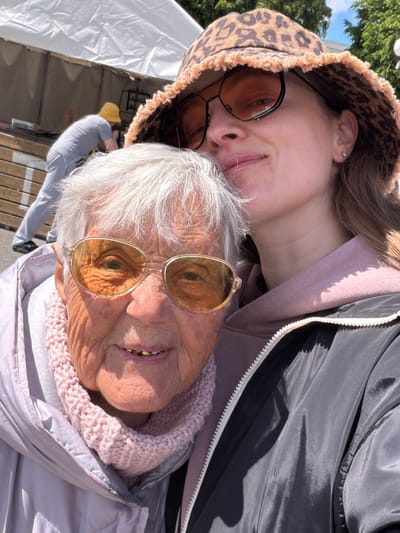The image shows two people posing closely together. The younger person, wearing a patterned bucket hat and tinted glasses, is positioned slightly behind the older person. The older person, also wearing tinted glasses, has white hair and a pink knitted scarf. A gray zippered jacket is visible. The background includes a white tent-like structure and a glimpse of a person in a gray shirt and yellow hat. The location, Valozhyn, Belarus, is not verifiable based on the image content alone.