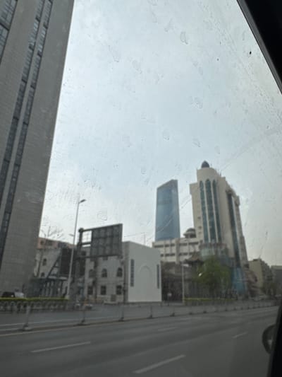 View from a window with smudges, revealing buildings and a street in Shenhe District, China. The buildings include a skyscraper and lower structures, while the street shows multiple lanes.