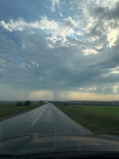This is an exterior, eye-level, vertical shot of a wet, two-lane asphalt road with a white dashed line in the center, curving to the right in the distance. The road is surrounded by green fields of grass and foliage. The sky is filled with dramatic, dark gray and white clouds, with shafts of sunlight breaking through in places. In the far distance, on the left side of the road, there are some trees and what appears to be a low-rise city or industrial area. On the right side, in the distance, there is a long, yellow-roofed building. The front of a vehicle is visible in the foreground, suggesting the image was taken from the driver's perspective. The environment appears to be rural, with evidence of recent rain. The city of Патруши, Russia, is mentioned as the location, though no specific landmarks within the city are definitively identifiable in the image.