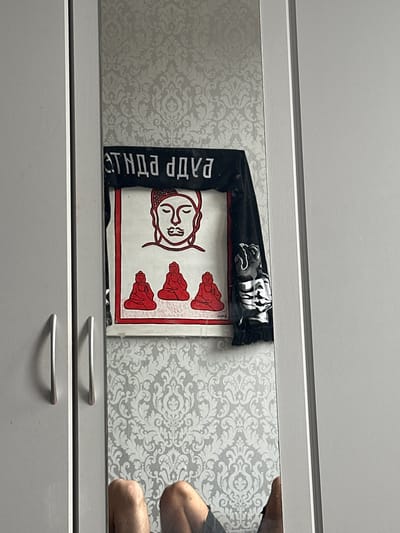 The image shows a partial view of a person's legs in the foreground, reflected in a mirror. Behind the reflection, a poster or artwork featuring red outlines of Buddha figures is visible. Above the artwork, there is a black banner with white text. The background of the mirror is decorated with a light-colored damask wallpaper. The scene appears to be indoors, possibly in a bedroom or living area. The photograph does not provide any specific information or context to indicate a location in the city of Hamm, Germany.