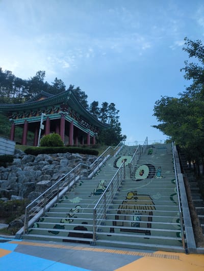 This image captures a wide-angle view of a traditional Korean pavilion, identified by the Korean text "천고각" (Cheongogak) on its facade, situated on an elevated, rocky embankment in Godang-ri, South Korea. The pavilion features a green-tiled, multi-tiered roof with ornate eaves, supported by numerous red pillars. Lush pine trees densely cover the hill behind the structure.

In the foreground, a prominent, wide staircase ascends towards the pavilion. The risers of these concrete stairs are painted with a series of colorful, cartoon-like illustrations depicting traditional Korean figures, drums, and cloud motifs, primarily in shades of green, yellow, and red on a light grey background. Polished silver-colored metal handrails with multiple horizontal bars line both sides of the staircase. To the far right, a narrower, undecorated set of stone stairs runs parallel to the main painted staircase.

The base of the hill beneath the pavilion is composed of various sizes of grey rocks, with a small, rounded green bush directly in front of the pavilion's center. The sky overhead is a clear, light blue with sparse, wispy white clouds scattered across it. On the right side of the image, the branches of a deciduous tree with green leaves are visible, extending into the frame. The ground at the bottom of the stairs features a geometric pattern of blue, yellow, and dark grey pavement sections, with the yellow areas incorporating tactile paving for accessibility. No individuals are present in the scene.