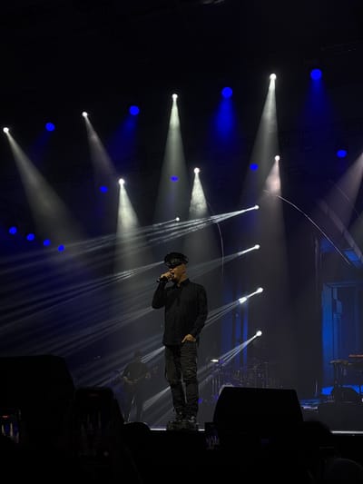 A performer stands on a stage illuminated by spotlights in Pagedangan, Indonesia. The performer is wearing dark clothing and holding a microphone. Behind the performer, there are other performers and musical instruments visible. Stage lighting in the form of white spotlights and blue stage lights are visible and directed toward the performers on the stage. Audio equipment and possibly audience members are visible in the darkness surrounding the stage.