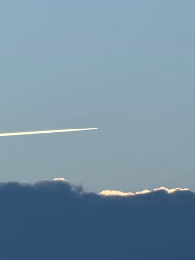 A contrail from an airplane stretches horizontally across a pale blue sky. Below the contrail, a thick, dark blue cloud band with a bright white, illuminated edge spans the lower portion of the frame. The scene is devoid of any identifiable city landmarks, people, or specific structures, and therefore cannot be geographically pinpointed to Палыкавіцкі сельскі Савет, Belarus, or any other specific urban location. The image captures a moment in the sky, featuring natural atmospheric phenomena.