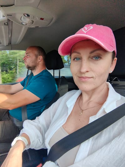 A medium shot shows a man and a woman in a car. The man, in the driver's seat, is wearing a teal t-shirt and has short, brown hair and a beard. The woman, in the passenger seat, is wearing a pink baseball cap with a white heart, a white button-up shirt, and a tan tank top. She has blue eyes and is looking towards the camera. A seatbelt is visible across her chest. The car's interior is visible, with the roof console overhead. The background shows greenery and trees, suggesting a rural or suburban setting, possibly on a road near Хаценчыцкі сельскі Савет, Belarus.