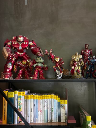 This media file captures a close-up view of a shelf filled with books and action figures. The dominant subjects are several Iron Man action figures, meticulously arranged, and a stack of books, some with colorful illustrations. The scene is set indoors, with a grey, textured wall on the left and the shelves on the right, suggesting a room or a shop.

The action figures appear to be in dynamic poses, suggesting a battle or dramatic scene, with Iron Man figures in various red and gold suits. One figure appears to be a Hulkbuster armor, with a smaller figure, possibly Hulk, positioned on top of it. The books are stacked vertically, with their spines facing outward. Many of the books have yellow labels with Thai numerals and text, indicating they are likely from Thailand. Some visible text on the book spines includes "นิ้วกลม" (Nichakorn), a Thai author, and titles like "กลางทะเลทราย" (In the Desert) and "สิ่งมหัศจรรย์ธรรมดา" (Ordinary Wonders). The bottom book has the text "THREE HOURS FROM".

The lighting suggests it is daytime, casting shadows and highlighting the details of the figures and book covers. The overall mood is one of display and collection, with a focus on the popular Marvel character, Iron Man, and a selection of Thai literature. The location context provided identifies the setting as Prachuap Khiri Khan, Thailand, which aligns with the presence of Thai text on the books. The image is rich in detail, showcasing a hobbyist's collection or a retail display.