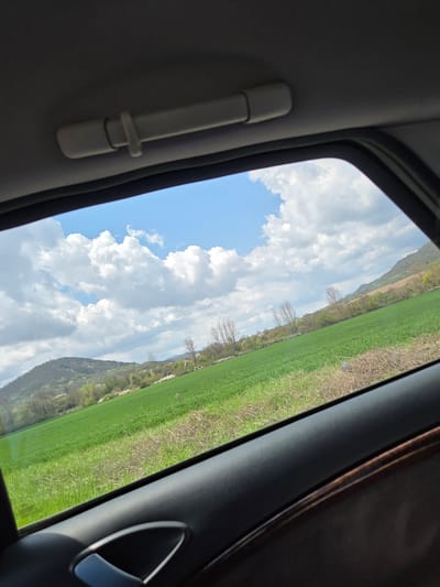 The image depicts a view from inside a vehicle, through a window. The view is of a landscape featuring a green field and a cloudy sky. A hill or low mountain is visible in the background. Parts of the car's interior are visible, including a grab handle and parts of the door or frame. The location is in Dobrodan, Bulgaria.