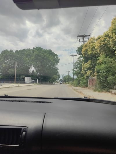 The image is taken from the interior of a vehicle, looking forward through the windshield. The view is of a two-lane asphalt road stretching into the distance under a cloudy sky. On the right side of the road, there is a dense line of green trees and bushes, with some lighter-colored flowering branches interspersed. Several utility poles with electrical wires run parallel to the road on the right. On the left side of the road, there are large, mature trees and some buildings, including what appears to be a white trailer or truck. The road appears to be in a semi-urban or suburban area. A signpost with a white circle and a directional arrow is visible on the left. A small, light-colored car is seen in the distance on the right side of the road. The foreground shows the dashboard of the vehicle. The overall impression is that of a slightly overcast day in a town or city in Greece, likely Komotini, given the context.