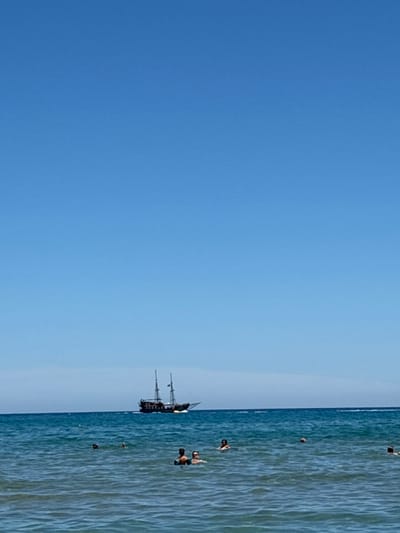 This is a photograph taken at the beach in Rethymno, Greece, under a clear blue sky. The sea is a vibrant blue, with moderate waves. In the distance, a sailing ship with multiple masts and dark sails is visible on the horizon. Closer to the shore, several people are swimming and enjoying the water. The focus is on a group of individuals in the foreground, with their heads and shoulders visible above the surface. The overall scene depicts a typical summer day at the beach.