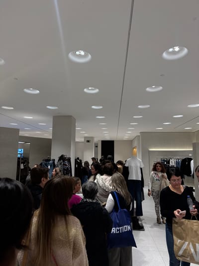 A long line of people waits in a modern retail store. The line extends into the depth of the frame. Most of the people are women. Some are carrying shopping bags. The store's interior is minimalist, with a white color scheme. The lighting consists of rows of recessed circular lights in the wall and ceiling. The architecture is boxy. Some clothing racks are visible along the back wall of the store.