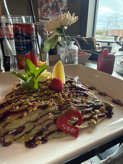 The image shows a plate of dessert in a restaurant or cafe setting. The main item is a crepe or pancake topped with a pistachio-colored sauce, drizzled with dark chocolate sauce, and sprinkled with chopped nuts, possibly pistachios. The dessert is garnished with a fresh raspberry, a slice of banana, a piece of white chocolate, and a sprig of mint. A red plastic heart with the word "LOVE" is placed on the side.
Beside the plate, there's a can of San Pellegrino Aranciata Rossa (Blood Orange) soda in a glass, along with other beverage bottles in the background. A small vase with a white flower is also present.
The scene is set near a window, providing a view of the outside, which includes trees and a glimpse of buildings across the street. Based on the presence of a "Geek Bar Pulse" vape device on the table, it appears the photo was taken in Willowbrook, United States of America.