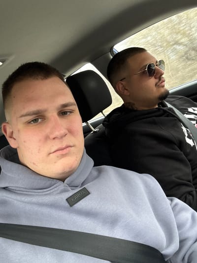 The scene depicts two adult males seated inside a vehicle, purportedly within or traveling through the city of Vetren, Bulgaria.
The subject in the foreground, positioned on the left, is a light-skinned male with a short buzz cut hairstyle and green eyes. He exhibits visible acne on his cheeks and jawline. He is wearing a light grey or periwinkle blue hooded sweatshirt featuring a black rectangular patch on the upper chest with the word "KARMA" in white capital letters. A black seatbelt is diagonally across his torso. His gaze is directed forward, seemingly towards the camera, with a neutral expression.
Behind and to the right of the foreground subject, the second male subject is visible. He possesses a darker skin tone, short dark hair with a fade, and a trimmed mustache and beard. He is wearing dark sunglasses with light gold-colored, hexagonal-framed lenses. A dark tattoo is partially visible on the right side of his neck. He wears a black hooded sweatshirt and a black seatbelt across his chest. His head is turned slightly to the right, and his gaze is directed away from the camera, looking towards the vehicle's right side or ahead.
The interior of the vehicle shows a light-colored headliner and a black headrest behind the foreground subject. Through the windows in the upper right quadrant of the image, a blurred outdoor background consistent with light green and brownish foliage or trees suggests the vehicle is in motion or parked outdoors.