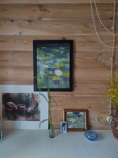 A still life composition is presented against a wooden paneled wall. The wall features visible wood grain and knots. On the wall, a framed watercolor painting of white water lilies on a pond hangs centrally. To the left of this painting, another framed artwork, a watercolor of purple onions, is placed lower on the wall. A small framed landscape painting with pink flowers sits to the right of the water lily painting, accompanied by a small, candid-style photograph with text below it. In front of these artworks, on a white surface, is a small green glass bottle with a single green sprig of foliage. To the right of the framed paintings, a light brown ceramic vase with a white decorative pattern holds a bunch of small yellow flowers. An electrical outlet with a light brown cover is visible on the wall to the right of the framed artwork. A glass dish with a lid, in a blue translucent color, sits on the white surface in the foreground. A beaded string and a rope with fasteners are also visible hanging from the top right of the frame, against the wooden wall. The scene is likely an indoor domestic setting.