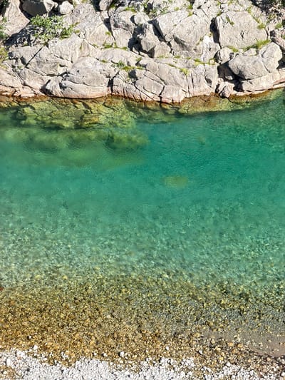 This overhead photograph captures a pristine natural environment in Unknown, Montenegro, showcasing a clear turquoise-green waterway alongside a rocky bank and pebbled shore.

The upper third of the image features a rugged, light-grey rock face composed of large, angular boulders with prominent fractures and crevices. Sparse green vegetation, consisting of small bushes and grass, grows intermittently from these rock formations. Direct sunlight from the upper left casts sharp shadows across the rocky terrain.

The central portion of the image is dominated by exceptionally clear, vibrant turquoise-green water. Near the rock face, the water is shallow, revealing submerged rocks and reflections of the cliff face with an olive-green tint. Further towards the center, the water appears deeper, maintaining its brilliant turquoise hue while still allowing visibility of the textured, pebbly bottom through subtle surface ripples.

The bottom third of the frame depicts the riverbed and a narrow strip of shoreline. Submerged pebbles, ranging in color from golden-brown to light grey, are distinctly visible through the shallow water, exhibiting a complex pattern. These gradually transition to an exposed, dry shoreline at the very bottom, characterized by a dense accumulation of predominantly white and light-grey pebbles. No individuals, man-made structures, or actions are present in the scene, which is illuminated by bright daylight.