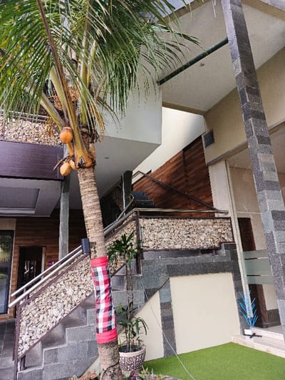 A low-angle shot showcases a tropical outdoor setting. A palm tree with coconuts dominates the left, wrapped in a red and white checkered cloth at its base. A staircase with stone and metal railings ascends to an upper level, featuring a wooden wall. Architectural elements include stone pillars and tiled steps. Potted plants add greenery at the base of the stairs. A grassy area is visible on the lower right.