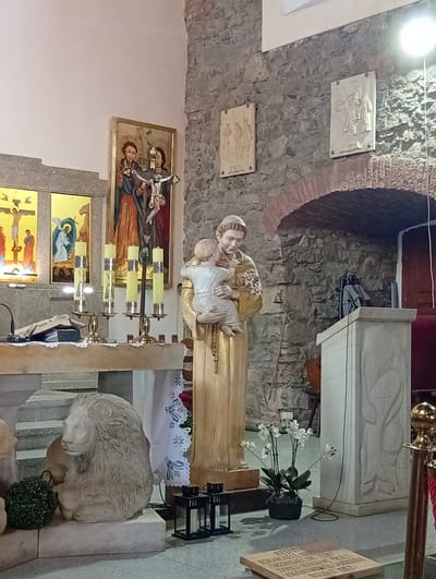 This is a photograph of the interior of the Church of St. Joseph the Betrothed in Sobótka, Poland. In the foreground, a stone sculpture of a lion is visible on the left. To the right of the lion is a large statue of Saint Anthony holding the infant Jesus. Behind the statue, several tall, yellow candles in brass holders are arranged on a stone altar. Above the altar, two religious paintings are displayed on the wall, depicting biblical scenes. The background features a rough stone wall with arched doorways and two carved relief panels mounted on the wall. A wooden podium with a carved relief is also present on the right side of the image. The lighting appears to be artificial, with a bright light source visible at the top right of the frame.