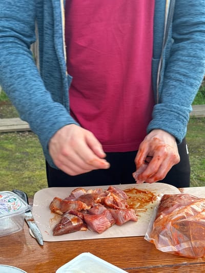 An outdoor scene, possibly in Maksimovo, Russia, features a person wearing a blue hooded jacket over a pink t-shirt. Their hands, stained with a reddish sauce, are actively engaged with raw meat. A cutting board displays several pieces of marinated meat, while a plastic bag on the right also contains marinated meat. A small plastic container and a knife are also present on a wooden table, set against a backdrop of greenery.
