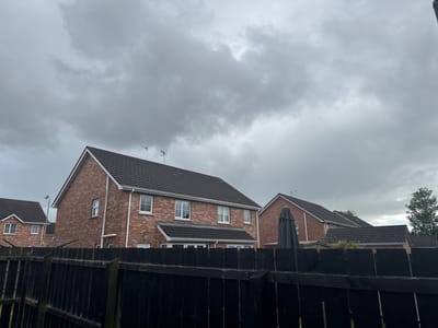 This is a medium-angle, full-shot photograph taken outdoors on an overcast day. The scene depicts a residential area with several brick houses visible. In the foreground, a black wooden fence runs horizontally across the frame, partially obscuring the view of the houses. The houses have dark tiled roofs and white-framed windows. Two of the houses have television antennas on their roofs. To the right, a large closed umbrella or parasol stands near one of the houses. A street lamp is visible in the distance behind the houses. The sky is filled with heavy, gray clouds, suggesting a cloudy or possibly rainy day. The overall atmosphere is subdued due to the weather and the lack of visible activity. The location is identified as Craigavon, United Kingdom. There is no visible text in the image.