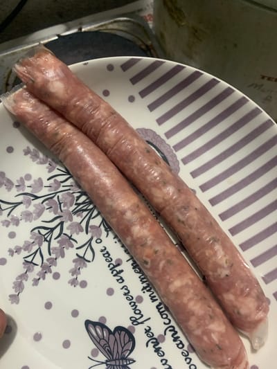 The image shows two raw sausages on a decorative plate. The sausages are elongated and appear to be wrapped in plastic. The plate is white with a pattern of purple stripes and lavender sprigs, along with small purple dots and a butterfly illustration. The sausages are positioned diagonally across the plate. The background is blurred but suggests a stovetop and possibly a pot or pan. The overall scene is likely an indoor setting, possibly a kitchen, given the presence of a stovetop and uncooked food. There is no indication of the city of Mechkarevo, Bulgaria, in the image.