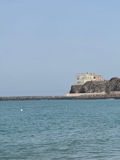 The image displays a serene coastal landscape scene, viewed horizontally, with the sky occupying the upper left portion, land and buildings in the center, and water to the lower right.
The scene features a rugged, dark gray rocky cliff face that dominates the midground. Several modern, multi-story white buildings with minimalist architecture, flat roofs, and balconies are constructed into and on top of this cliff. These structures appear to be residential or resort-style, blending into the natural rock.
A robust retaining wall or breakwater, constructed from large rocks and concrete, runs along the base of the cliff, separating it from the expanse of water. The water, a calm turquoise-blue, shows subtle ripples and texture across its surface. Two small, circular white buoys are visible floating on the water, suggesting navigation or marking a boundary.
The sky is a uniform, clear light blue-gray, indicating daytime, possibly early morning or late afternoon given the soft, diffused light. There are no people visible, and no specific activity is taking place, contributing to a tranquil and peaceful ambiance.
Faint, dark text or numbers are vaguely discernible on a section of the rock face behind the buildings, but they are too indistinct to read clearly. A small sign on the retaining wall is also present but illegible. The overall setting suggests a modern coastal development harmoniously integrated with the natural, rocky landscape, consistent with locations like Al Fiqait in the United Arab Emirates.