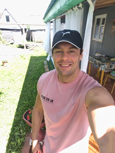 A man wearing a pink sleeveless t-shirt and a black baseball cap with a white Puma logo smiles at the camera in a selfie. He is standing outdoors on a sunny day, with a green lawn, a flower pot with red flowers, and some wooden structures in the background. A covered outdoor seating area with white curtains and a table with food is visible to his right. The man appears happy and relaxed. The image seems to have been taken in the afternoon, given the strong sunlight and shadows. The text on his t-shirt reads "ES ES ES IS" in a vertical, blocky font. The location is Yaganovka, Russia.