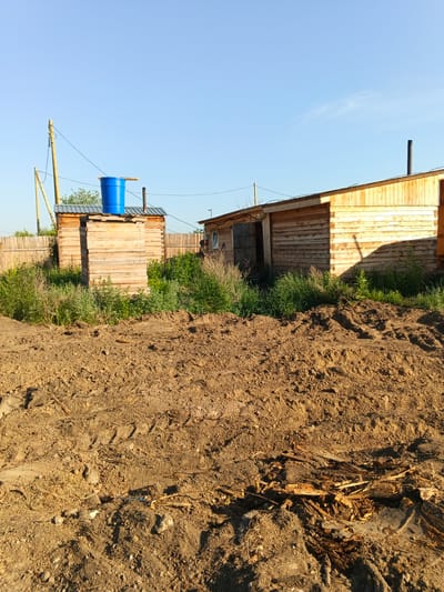 In Strelka, Russia, a rural setting is depicted with two wooden structures under a clear blue sky. In the foreground, a rough, uneven dirt area with visible tire tracks suggests recent activity or construction. To the left, a tall, slender wooden outhouse stands with a bright blue barrel perched on its roof. Behind the outhouse, a wooden fence runs horizontally. To the right, a longer, low-slung wooden building with a corrugated metal roof is visible. This building has an open doorway revealing darkness within and a dark chimney pipe protruding from its roof. Overgrown green vegetation surrounds the base of both structures. The sunlight casts distinct shadows on the wooden buildings.