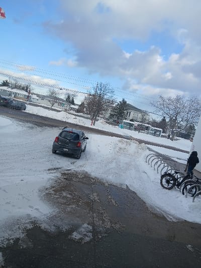 A tilted outdoor scene shows a street partially covered in snow. A dark-colored car is parked on the snowy street. Bicycles are secured in a rack nearby. A person stands beside the bikes. Houses line the opposite side of the street. The sky is predominantly blue with scattered clouds. Overhead power lines are visible.