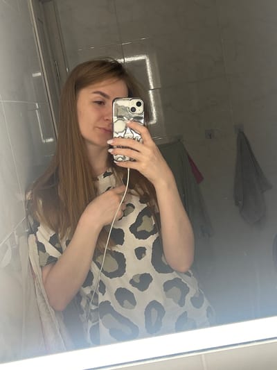 A young Caucasian woman with long brown hair is taking a selfie in a bathroom. She is wearing a light-colored t-shirt with a leopard print pattern. Her right arm is extended, holding a smartphone with a silver skull-patterned case. A white cord is visible extending from the phone. The background is a tiled bathroom wall with a large mirror reflecting the scene. Two towels are hanging on the wall behind her. The lighting suggests an artificial light source above the mirror. The location is the bathroom of a residential dwelling in Нижние Муллы, Russia.