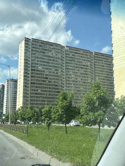 The image, originally rotated 90 degrees clockwise, captures an urban or suburban street scene on a bright, cloudy day, seemingly from inside a vehicle. On the left, two prominent, tall multi-story buildings dominate the skyline, featuring a grid-like facade of numerous uniform windows, suggesting residential or office use. A third, similar building is visible further down the street. The buildings stand against a blue sky dotted with scattered white clouds.

To the right, a wide strip of vibrant green grass is punctuated by several lush, green deciduous trees. A paved road with a clear curb runs alongside the grassy area. A white car is partially visible behind trees, and another white car can be seen further down the road. Infrastructure includes dark power lines stretching across the upper left, a street light pole in the lower right, and a circular manhole cover on the road. A low, metal fence borders a section of the grassy area. The diagonal lines of a car's interior or window frame are visible along the top and far right edges, indicating the vantage point. It is daytime with ample natural light. No people or specific activities are discernible. There is no visible text. The architectural style and urban setting are consistent with areas in Russia, matching the provided context of Poroshkino.
