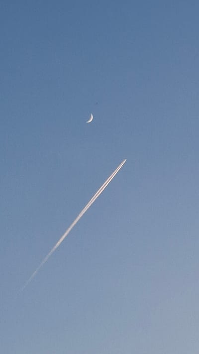 The image depicts a bright blue sky with two primary subjects: a crescent moon and a contrail formed by an airplane. The moon is located on the left side of the frame, appearing as a thin, curved shape. The contrail stretches diagonally across the sky from the upper left to the lower right, suggesting the path of a jet. The contrail gradually dissipates toward the lower right. There are no people or other objects visible. The image was taken in the city of Шешурга, Russia.