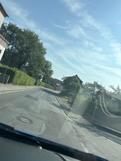 The image depicts a view from a vehicle, looking out onto a two-lane asphalt road in a residential area, likely in Volpertshausen, Germany. The road curves slightly to the left, with a raised pavement running along the right side. A speed limit sign indicating 30 km/h is visible on the right, near a streetlamp. Lush green hedges and trees line the sides of the road, with houses visible in the background. The sky is bright blue with scattered white clouds. A manhole cover is present in the road surface on the left. The overall scene suggests a calm, daytime setting in a suburban environment.