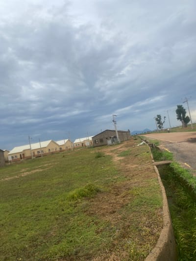 The image captures a partially constructed residential area in Bulamare, Nigeria. The scene is viewed from a low angle, looking upwards towards a cloudy sky. In the foreground, a grassy area with patches of dry earth slopes away, bordered by a concrete curb and a paved road with some overgrown vegetation along its edge.
Several newly built houses are visible in a line, their pale exteriors and white roofs contrasting with the natural surroundings. Some of the houses appear to be in the early stages of construction or finishing. Electrical poles and wires are scattered throughout the scene, indicating the presence of infrastructure. In the distance, silhouetted trees and what appears to be a mountain range can be seen against the sky, suggesting a rural or semi-rural setting. The lighting and cloudy sky suggest it is daytime, possibly late afternoon, with overcast weather. There is no discernible activity or people in the image. No text is visible.
