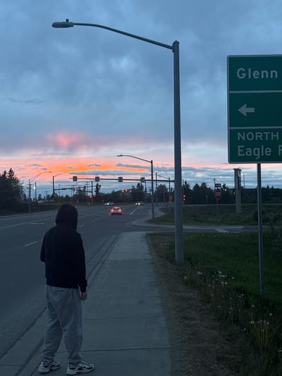 The image depicts a street scene at dusk in Anchorage, Alaska. A person wearing a black hooded sweatshirt and grey sweatpants stands on a sidewalk, facing away from the camera and towards a road. The sky is filled with colorful clouds, displaying hues of orange, pink, and blue, indicating either sunrise or sunset. A street light looms over the sidewalk. Across the road, traffic lights are visible, and several cars are driving. A green directional sign with white lettering is mounted on a pole, indicating "Glenn" with an arrow pointing left and "NORTH Eagle P" below. A stop sign is also visible further down the road. The environment is characterized by asphalt roads, sidewalks, grassy areas with dandelions, and trees.