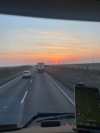 The image depicts an eastbound view from inside a moving vehicle on a multi-lane road near Slaný, Czechia, at approximately 06:37 AM during sunrise. The sky transitions from light blue at the zenith to vibrant orange and red at the horizon, where a distinct red solar disk is visible.
The paved road features two visible lanes in the direction of travel, separated by dashed white lines, with a solid white line marking the right shoulder. A white box truck occupies the right lane in the mid-distance, followed by a silver sedan with visible license plate "6U0 9940" in the left lane, both traveling away from the observer. The surrounding terrain consists of flat, open fields or grasslands, appearing sparsely vegetated. Utility poles carrying power lines are visible along the right side of the road.
In the foreground, the top of the observer's vehicle dashboard and windshield wipers are partially visible. A mobile device, likely a smartphone or tablet, is mounted on the dashboard to the right, displaying a navigation map. The navigation interface indicates "Praha" (Prague), a distance of "17 km," a speed limit of "80," and the current time as "6:37." It also displays "14 km" to "Bosna Žižice," a locality in the immediate vicinity of Slaný, Czechia.