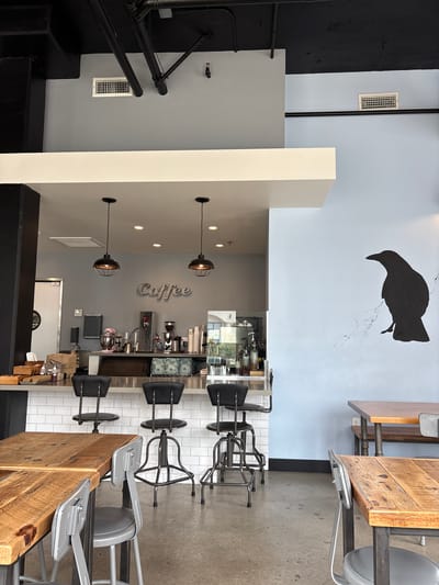 The image depicts the interior of a modern coffee shop. The foreground features two rustic wooden tables with metal chairs. Behind the tables is a counter with three swivel stools. The counter area has a tiled backsplash and displays various coffee-making equipment, including espresso machines, a grinder, and a pink stand mixer. The word "Coffee" is spelled out in raised lettering on the wall behind the counter. To the right of the counter, a large black silhouette of a bird is painted on a light blue wall. In the background, there are more tables and chairs, suggesting a seating area. The ceiling is black with exposed pipes and black lighting fixtures. The overall atmosphere is clean and contemporary. This coffee shop is located in Long Beach, California, United States of America. There are no people visible in the image.