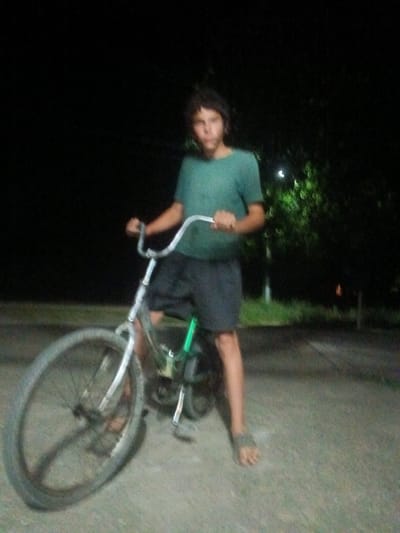In Leontyevskoye, Russia, a person is standing next to a bicycle at night. The person, who appears to be a young male with dark, curly hair, is wearing a teal t-shirt and dark shorts. He is positioned on the left side of the bicycle, with his left foot resting on the ground. The bicycle is a silver-colored frame with black tires and a green component visible on the rear wheel assembly. The background consists of dark foliage and a blurred light source, suggesting an outdoor, possibly rural or suburban, setting illuminated by artificial light. The ground appears to be paved or dirt.
