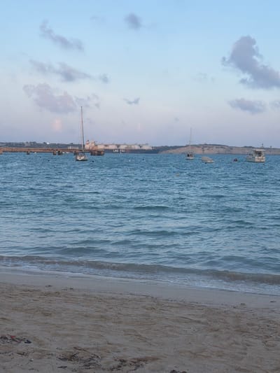The image depicts a coastal scene in Birżebbuġa, Malta, overlooking Marsaxlokk Bay. In the foreground, a sandy beach occupies the lower third of the frame, characterized by light brown sand and minimal scattered debris. Small waves with white foam crests gently break on the shore.
The midground is dominated by the blue-green sea, exhibiting a ripple pattern across its surface. Anchored within this area are three boats: a white sailboat with a visible mast positioned slightly left of center, a smaller white boat partially covered by a light-colored tarp to its right, and a white motorboat with a cabin situated further right towards the distant coastline. Multiple small yellow buoys are dispersed across the water, indicating mooring locations.
In the background, a long, dark-colored pier or jetty extends horizontally from the left side of the frame, supported by multiple pilings. Beyond the pier and anchored in the bay is a large, dark-hulled vessel identified by "LNG FSU" text on its side, a Liquefied Natural Gas Floating Storage Unit, featuring several large, spherical storage tanks on its deck. The distant right background shows a light-colored, elevated landmass, likely a section of the coastline. The sky above is a light blue with scattered white and light grey clouds. No human subjects are visible.