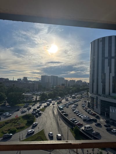 The image depicts an aerial view of a cityscape in Tarasovka, Russia, likely during daylight. The scene is dominated by a modern, multi-story building with a reflective facade, a parking lot filled with numerous cars, and a winding road with traffic. The sun is visible in the sky, casting a bright light and creating a dynamic atmosphere with scattered clouds.
Several cars are parked in rows in the lot, while others are on the road, indicating ongoing vehicular activity. The architecture suggests an urban or suburban setting. There are trees and greenery interspersed throughout the scene, providing a contrast to the built environment. The overall mood is one of everyday urban life, with the bright sunlight suggesting a pleasant day.
There are no clearly discernible people in the image. The visible text on the modern building is in Russian and appears to be signage for businesses, although it is not sharp enough to be fully legible. The time of day is likely late morning or early afternoon, given the sun's position and brightness. The weather appears clear with some cloud cover.