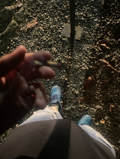 A person stands on small gravel, holding a lit cigarette. Light-wash, ripped jeans and blue running shoes are visible. A black item of clothing is also present. Some dry leaves are scattered on the gravel. A metal post with attached brackets is on the left side of the image.
