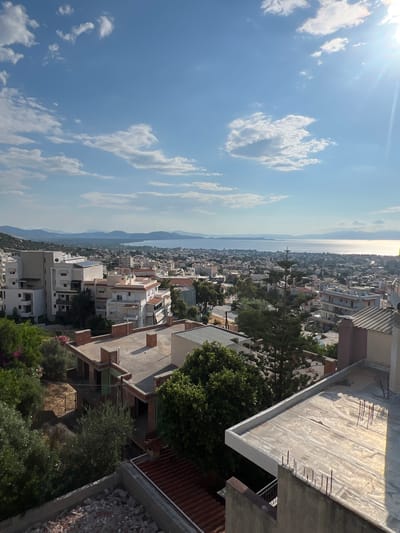 This is a high-angle, sunlit, outdoor, landscape photograph taken in the municipal unit of Nea Makri, Greece. The scene captures a panoramic view of a coastal town nestled between hills and the sea under a bright, partly cloudy sky.
In the foreground, the tops of several buildings are visible. One prominent structure is a building under construction with exposed brickwork and concrete foundations. Another building to its right has a flat, light-colored roof with rebar protruding from the concrete. Further back, a dense cluster of residential buildings with various architectural styles, including apartment blocks and houses, spreads across the landscape. Many buildings are white or light-colored, with red-tiled or flat roofs. Some buildings feature balconies and solar panels.
Greenery is scattered throughout the scene, with trees and bushes visible around the buildings. A tall, coniferous tree stands out in the middle ground.
The middle and background are dominated by the expansive, calm sea, reflecting the sunlight. Distant, hazy hills form a gentle horizon line across the water. The sky is a vivid blue with scattered white clouds, and the sun is bright, casting a lens flare in the upper right portion of the image. The overall impression is of a sunny day in a Mediterranean coastal town. There are no people or animals visible in the photograph.