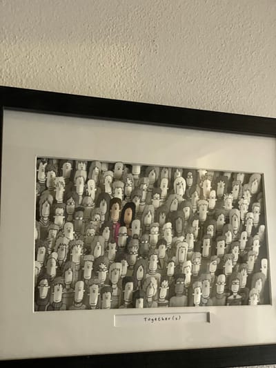 A framed piece of art hangs on a white textured wall. The artwork depicts a densely packed crowd of grayscale figures, each seemingly an individual, drawn in a cartoonish style. The figures are mostly facing forward, and their faces are minimalist in detail. Amidst the gray figures, there are two figures colored in, one with brown hair and the other with pink. A white label on the right side of the frame reads "Together (2)". The frame itself is black, and inside, the artwork is surrounded by a white matte border. The location is Clearwater, United States of America.