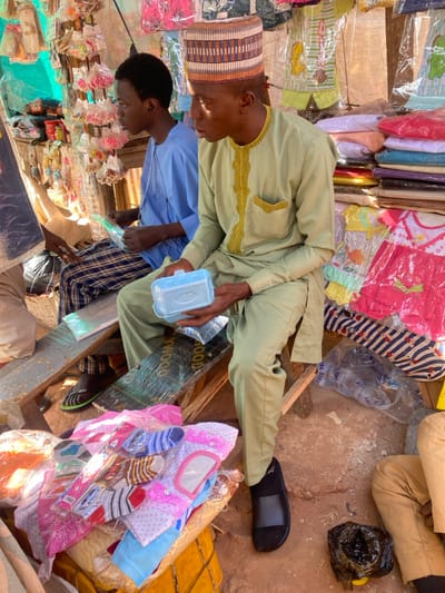 The scene depicts a market stall in Sabon Gari South, Nigeria, with two adult males seated on makeshift wooden benches. The man in the foreground, right, wears a light green traditional tunic with gold embroidery, matching trousers, and a striped multi-color cap. He holds a light blue plastic container in his left hand and a small clear plastic-wrapped item in his right. He wears black slide sandals. The man in the background, left, wears a light blue traditional top and plaid trousers. He holds a clear plastic-wrapped item, possibly stationery, and has an earbud in his left ear. He wears green flip-flops. Behind the men and to their right, various merchandise is displayed, including children's clothing (pajamas, dresses) hanging in clear plastic bags, stacked folded garments, and small packaged items. In the lower left foreground, a pile of children's socks and small garments, some individually packaged, rests on what appears to be a display. The ground is reddish-brown earth. A partial view of a third individual's lower legs and hand is visible in the lower right.