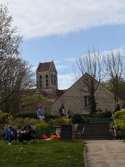 This image shows a church bell tower in Auvers-sur-Oise, France, with people sitting on chairs in a grassy area in the foreground. The sky is partially cloudy. There is a two-story building adjacent to the church with a sign that reads: "On adore les Pique-niques... Mais pas sur la Tarasse du Resto merci de m'éviter une Crise de Nerfs." There are bare trees and other greenery scattered throughout the scene.