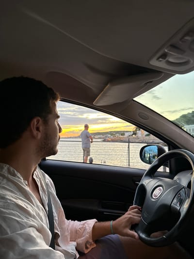 An image captured from within a parked vehicle, facing forward and slightly left, depicts a coastal scene in Nardò, Italy, during late afternoon or early evening, characterized by sunset colors.

In the foreground, the upper torso of a male driver, approximately 20-35 years old, is visible from the chest up, positioned in the driver's seat. He has short dark hair and stubble, wears a light-colored, unbuttoned collared shirt and dark shorts. A turquoise beaded bracelet is on his right wrist. His right hand rests on a black Fiat brand steering wheel. He is looking out the driver's side window towards the left horizon. The vehicle's interior features a dark dashboard and door panel, with a light-colored headliner and sun visor overhead. The driver's side mirror is visible, reflecting ambient light and distant objects.

Outside the vehicle, a coastal waterfront is visible. A dark metal barrier or railing runs parallel to the vehicle, separating a body of water from the foreground. On the water's edge, partially obscured by the railing, a dark, spherical object (possibly a buoy or mooring ball) is present. Beyond the railing, a male individual, appearing middle-aged to elderly, stands with his back to the camera, looking out over the water. He has short, light-colored hair and wears a light-colored t-shirt and light-colored shorts.

The body of water extends to the horizon, where the sky exhibits a prominent gradient of warm sunset hues, from bright orange and yellow directly at the horizon to lighter blues and greys overhead. On the opposite side of the water, distant white buildings of a coastal town are visible along a sloped landscape. The scene is illuminated by the low-angled light of the setting sun.