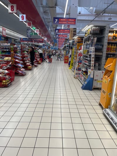 This image shows the interior of a large supermarket, likely a "Superstore" or similar hypermarket, given the scale of the aisles and the variety of goods on display. The floor is tiled with a white grid pattern. The ceiling is high, with white panels and visible light fixtures. In the distance, aisles are stocked with a wide range of products including food items, household goods, and possibly stationery or craft supplies based on the visible packaging and displays.

Several shoppers are visible throughout the store. On the left side, a person in a black t-shirt and jeans is standing, possibly looking at products. Further down, a man and woman with a shopping cart are visible. On the right side, several people are browsing shelves. A prominent orange display stand for "BIC" products is visible on the right side of the image.

Various signage is present, including directional signs, product category labels, and promotional banners. Numbers are displayed on hanging signs, such as "8", "9", "10", "11", and "12", which likely indicate aisle numbers or sections within the store. One sign with green arrows indicates an emergency exit. Other signs visible include "BISCOTTI", "FROLLINI", "PASTICCERIA", "LEGUMI", and "TOILETTI".

The overall impression is of a busy, well-stocked supermarket. Without further information or distinct landmarks, pinpointing the exact location within Castiglione delle Stiviere is not possible based solely on the image. However, the store appears to be a typical large-format retail establishment.