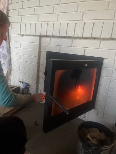 The image captures an indoor scene focused on a fireplace insert with a glowing fire visible through its glass door. A person's hands are visible on the right side of the frame, seemingly interacting with a control mechanism or latch on the fireplace. Their right hand is reaching towards the insert, while their left arm, wearing a teal long-sleeved shirt, extends across the frame. The fireplace is set into a white brick wall, with a visible texture suggesting painted or whitewashed bricks. To the right of the fireplace, a black bucket filled with what appears to be wood or kindling is partially visible.
The overall lighting suggests an indoor setting, likely in the evening or a dimly lit room, given the prominent glow from the fire. The perspective is from below, looking upwards towards the fireplace. The scene is set in Klepiki, Russia, as indicated by the location context. There is no visible text in the image. The emotion conveyed is one of quiet activity, perhaps tending to the fire.