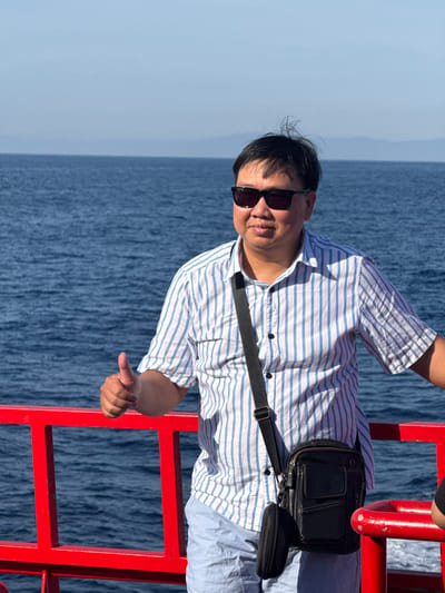 A man stands on the deck of a boat, giving a thumbs-up gesture. He is wearing a blue and white striped shirt, light blue shorts, and sunglasses. A black crossbody bag is slung over his shoulder. The boat has a red railing. The background consists of the blue ocean and a hazy horizon. The location is identified as Dong Son, Vietnam.