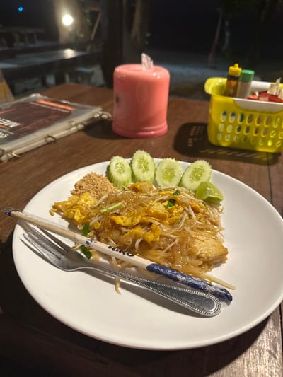 The image depicts a plate of Pad Thai served at a nighttime outdoor dining establishment in Phra Ae, Thailand. The dish, presented on a white circular plate, consists of stir-fried rice noodles with egg, bean sprouts, chopped scallions, and what appears to be chicken or tofu. It is garnished with a wedge of lime, a pile of ground peanuts, and five slices of fresh cucumber. A silver fork and a pair of white chopsticks with blue patterned tips and the brand "RIKO" printed on them rest diagonally across the plate.
The plate sits on a dark brown wooden table, which shows visible grain and texture. To the left of the plate, a menu in a clear plastic sleeve is partially visible, folded open. Behind the menu and to the right, a pink cylindrical object, possibly a decorative LED lamp or candle, emits a soft glow. Further to the right, a yellow plastic caddy with a perforated design holds small bottles of condiments (e.g., sauces, spices) and what appear to be small red fruit pieces (possibly strawberries or chillies).
The background is dimly lit, indicating an evening or nighttime setting. Blurred shapes of additional wooden tables and chairs are discernible in the distance, suggesting an open-air restaurant or a similar dining area. A faint light source is visible in the far background. No people are directly visible in the frame.